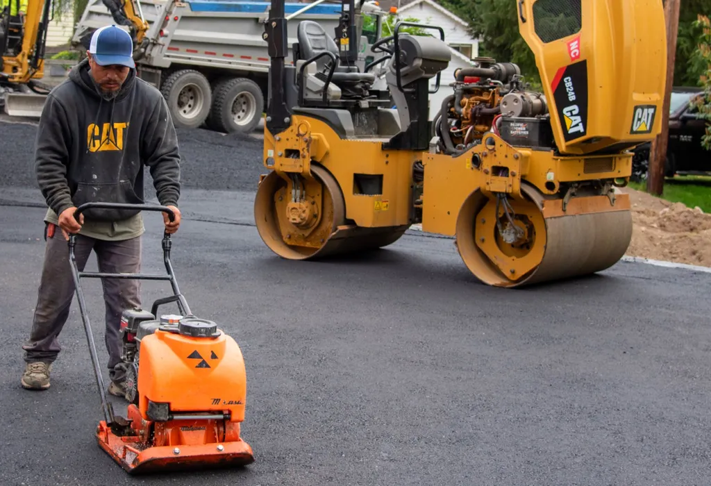 Crew member running a plate compactor across fresh asphalt beside a CAT roller
