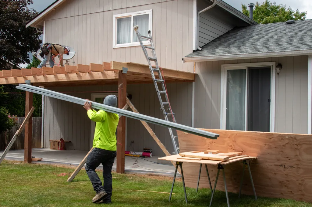 Crew framing a patio cover on a two story home while one worker carries a steel beam across the lawn