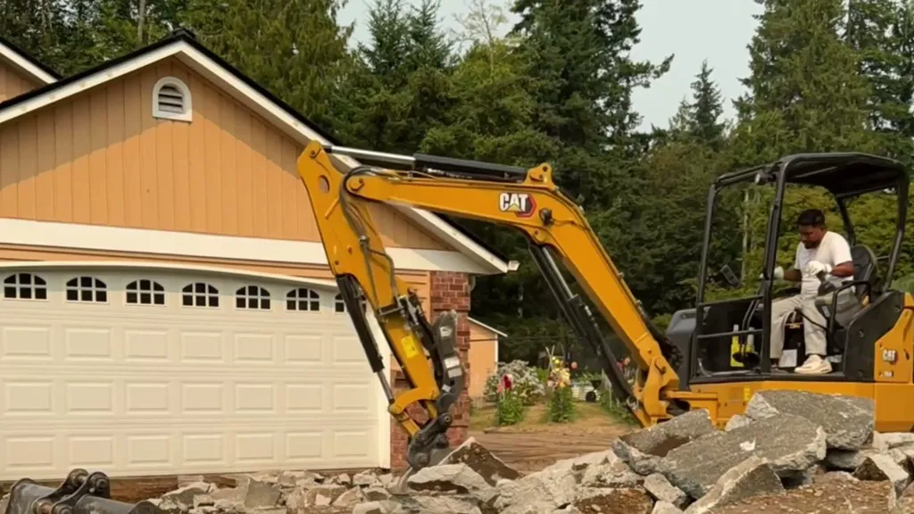 Operator in a CAT mini excavator breaking out an old concrete driveway in front of a home garage
