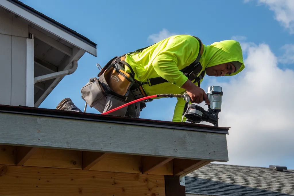 Worker in a high visibility hoodie and safety harness nailing shingles onto a new patio cover roof