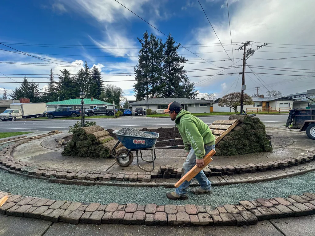 Crew member carrying lumber past curved brick paver borders, rolls of sod, and a wheelbarrow of gravel in a front yard