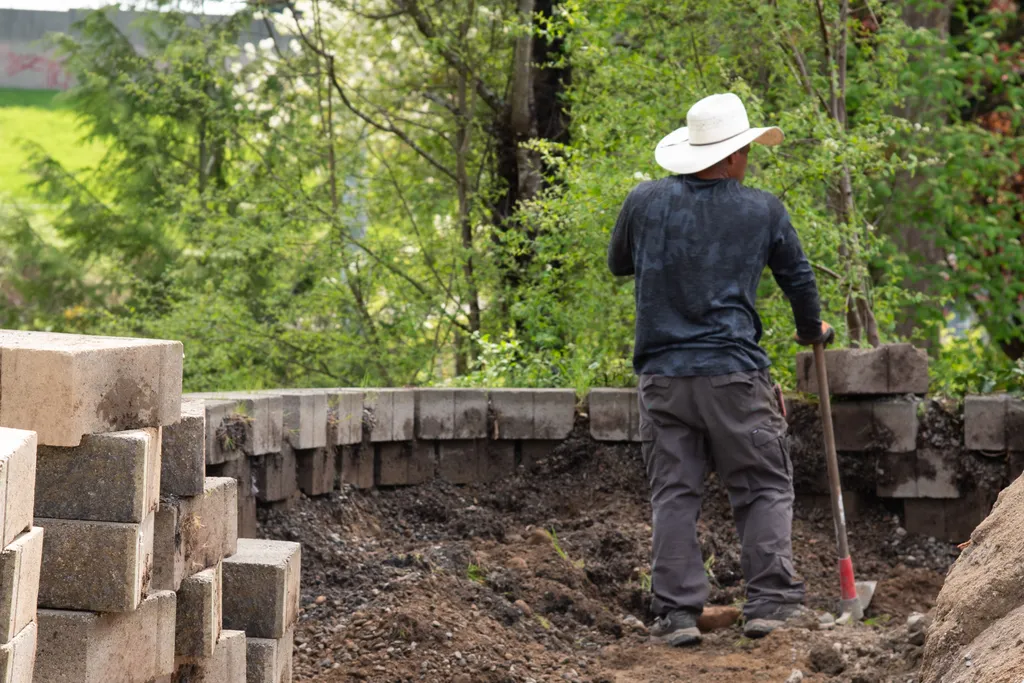 Worker with a shovel backfilling behind a curved block retaining wall, with stacked wall blocks in the foreground