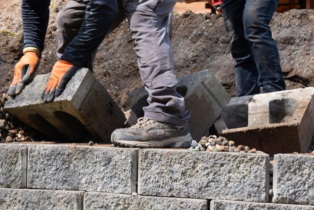 Workers setting concrete retaining wall blocks onto a course of wall block over a gravel base