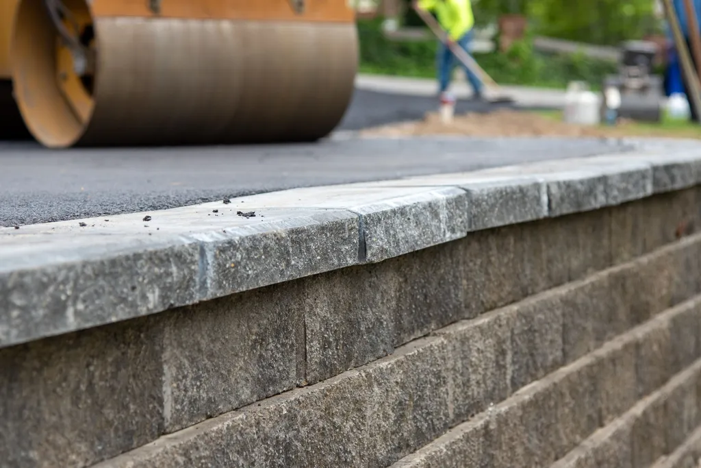 Close view of a capped block retaining wall along a freshly paved asphalt driveway with a roller behind it