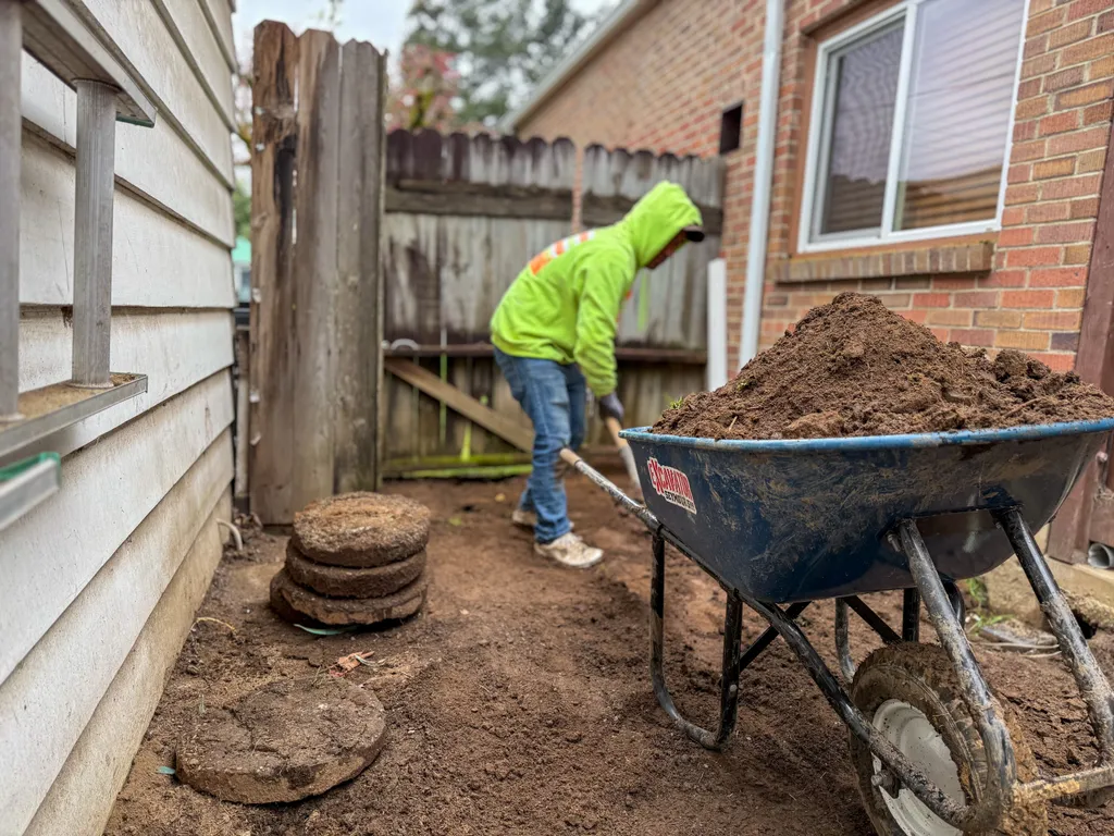 Worker grading soil in a narrow side yard next to a loaded wheelbarrow and a stack of stepping stones