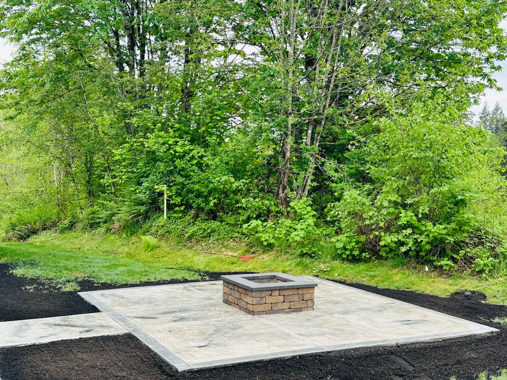 Square stamped concrete patio with a stone block fire pit, framed by fresh mulch at the edge of a wooded yard
