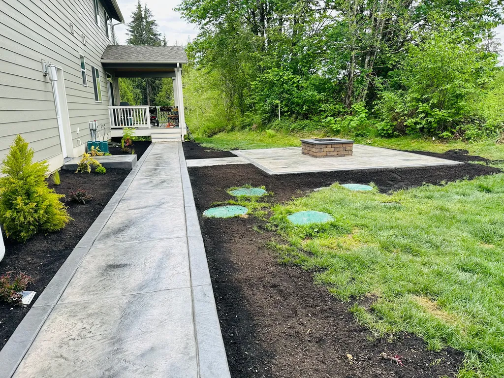 Stamped concrete walkway with a darker border running past new mulch beds and lawn to a fire pit patio